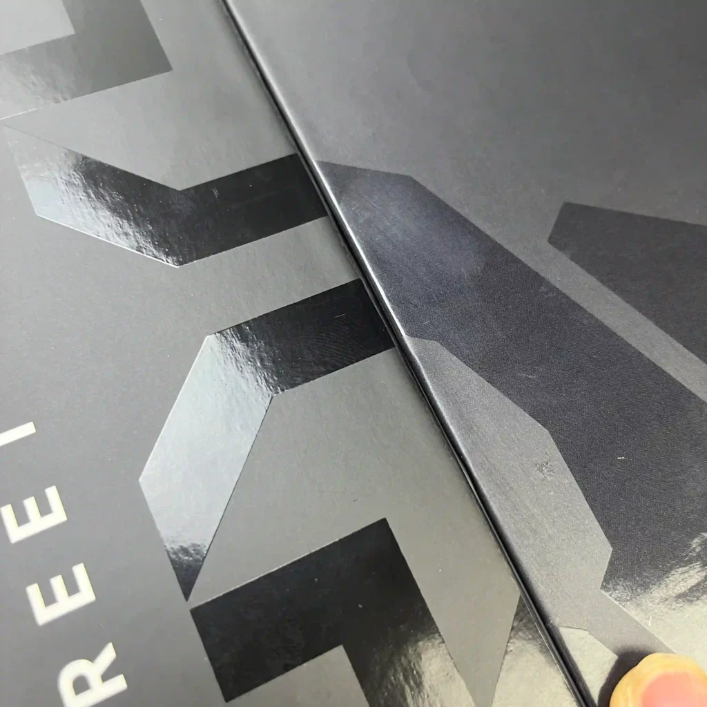 Comparison of Blind Spot UV and Black Colored UV on a matte black rigid box. The high-gloss varnish creates a subtle peek-a-boo logo effect against the matte lamination, while the colored UV provides a bolder, high-contrast finish for luxury branding